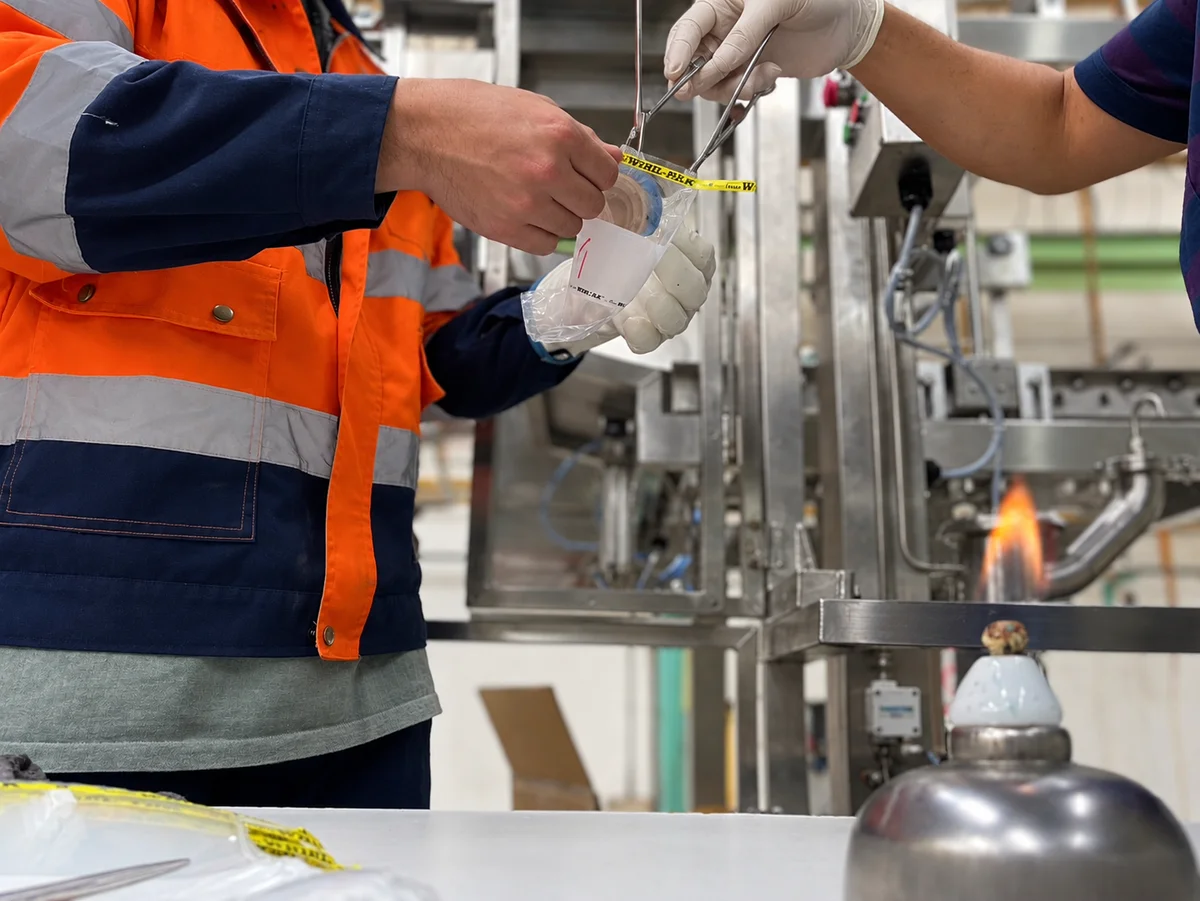 Operators handle a sample bag with tools near filling equipment, with a small flame and equipment in the foreground.