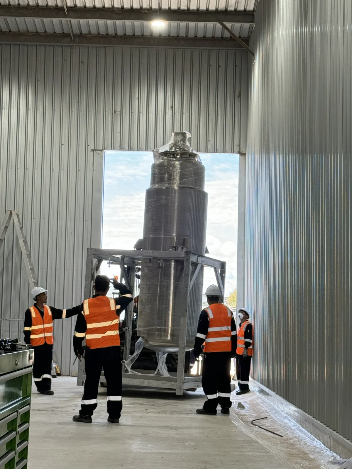 Personnel positioning a large vertical stainless-steel vessel inside a building.