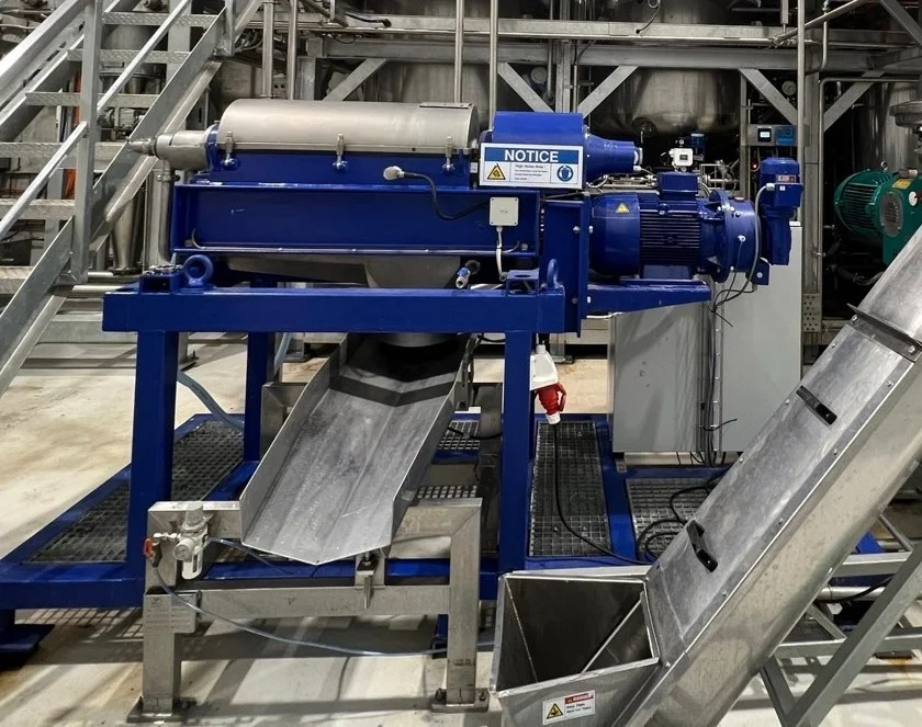 Blue horizontal decanter assembly with a motor, stainless-steel cover and discharge chute below.